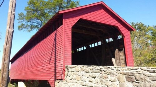 A red covered bridge with a stone foundation, surrounded by trees and a clear blue sky.