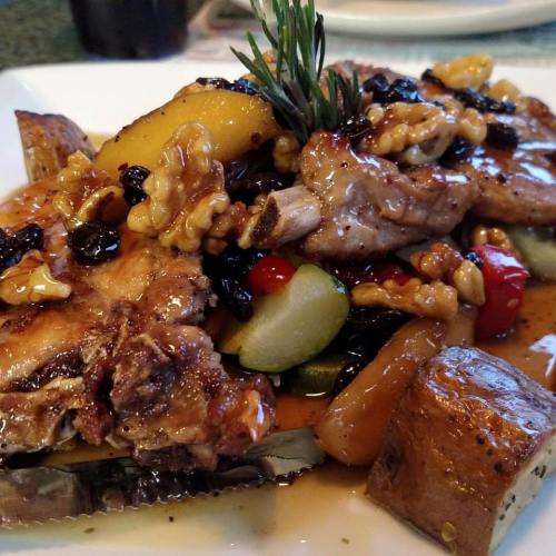 A plate of grilled meat topped with nuts and fruits, served with colorful vegetables and a drizzle of sauce.