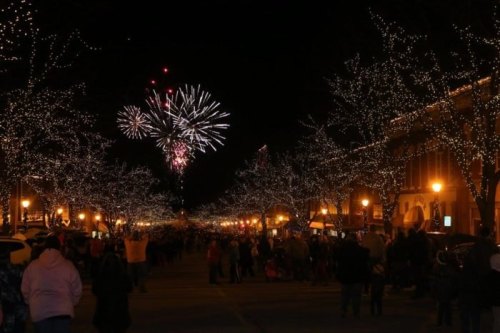 A festive street illuminated by lights, with a crowd watching fireworks in the night sky.