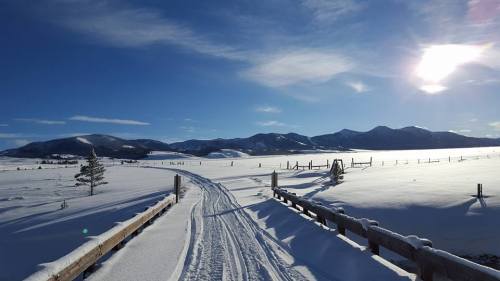 A snowy landscape with a winding path leading through fields and mountains under a clear blue sky.