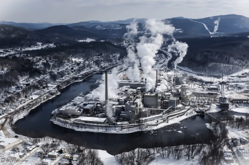 Aerial view of a snowy industrial area with smoke rising from factories near a river, surrounded by mountains.