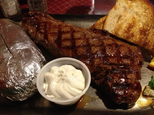 A grilled steak on a plate with a baked potato wrapped in foil and a side of sour cream, accompanied by toasted bread.