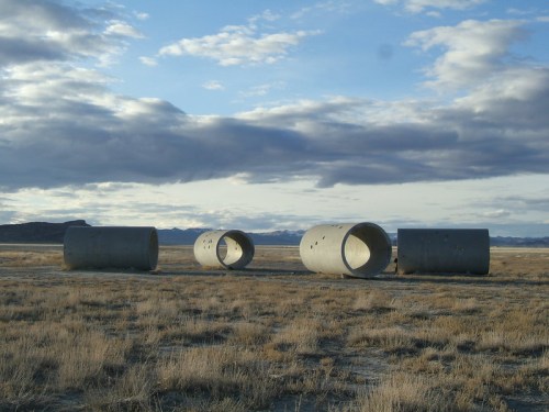 Concrete pipes scattered across a dry, grassy landscape under a cloudy sky with distant mountains.