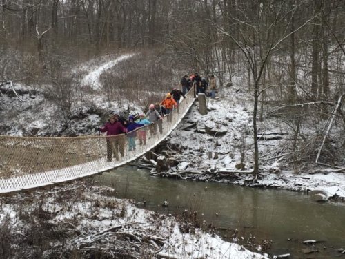 A group of people crossing a snowy suspension bridge over a creek in a wooded area.