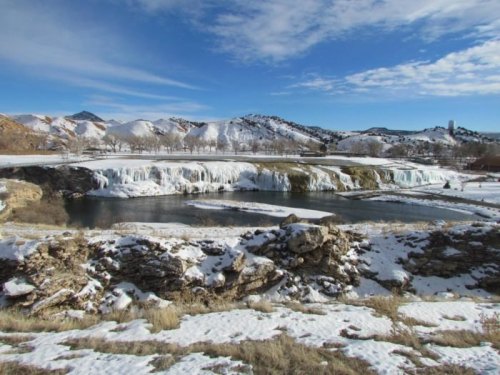 A snowy landscape featuring a frozen waterfall, a river, and mountains under a clear blue sky.