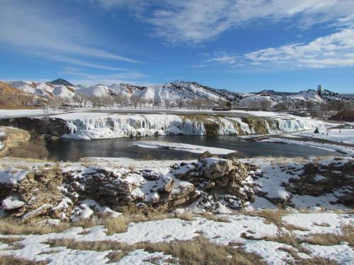 A frozen waterfall cascades into a river, surrounded by snow-covered land and mountains under a clear blue sky.