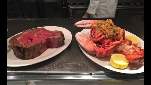 A platter with a large cut of prime rib and a seafood dish featuring lobster and shrimp, garnished with lemon.