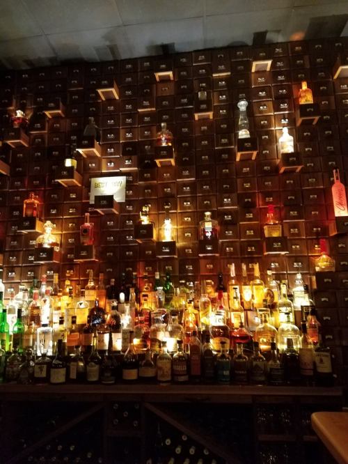 A well-lit bar shelf filled with various bottles of liquor, arranged in wooden cubbies against a dark wall.