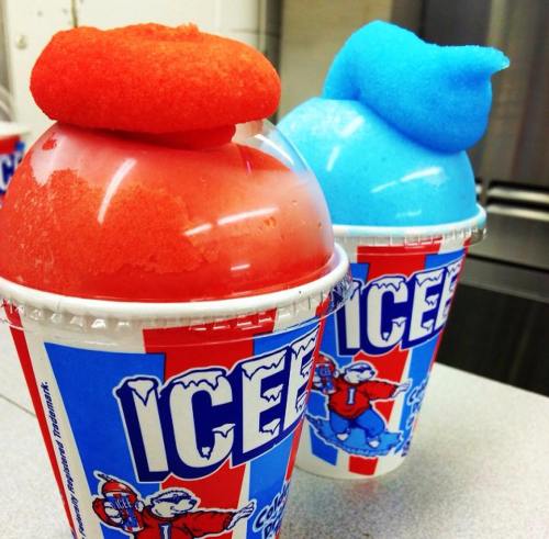 Two colorful ICEE cups, one with red and the other with blue slushy, topped with fluffy foam.