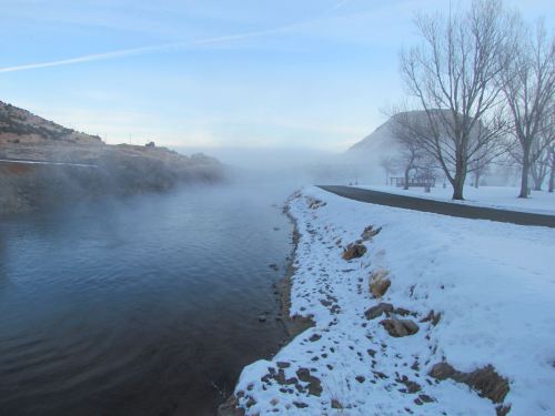 A misty river scene with snow-covered banks and bare trees, set against a backdrop of hills under a blue sky.