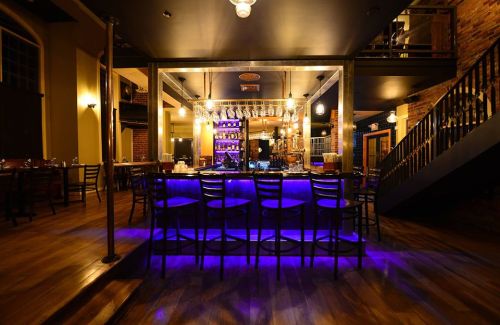 A stylish bar with purple lighting, wooden floors, and a modern design, featuring empty chairs and shelves of drinks.