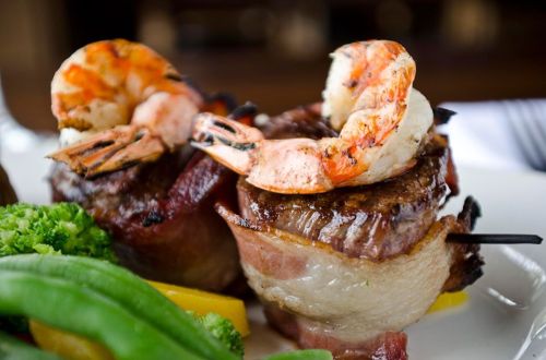 Grilled shrimp atop bacon-wrapped beef, served with broccoli and yellow bell pepper on a white plate.