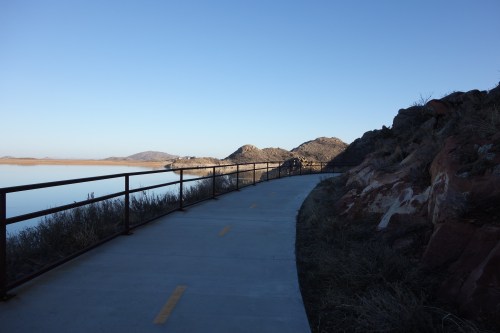 A paved path along a calm lake, bordered by a metal railing and rocky hills under a clear blue sky.