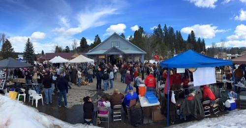 A bustling outdoor market scene with crowds, tents, and a blue sky, surrounded by trees and mountains.
