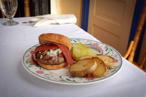 A plate with a burger topped with salami, pickles, and roasted potato slices, set on a white tablecloth.