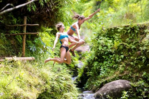Two girls in swimwear joyfully jumping into a lush green stream surrounded by rocks and foliage.