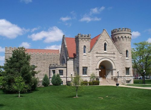 A stone building with a castle-like design, featuring turrets and a red-tiled roof, surrounded by green grass and trees.