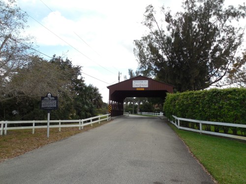 A covered bridge entrance with a sign, surrounded by trees and a white fence, leading to a road.