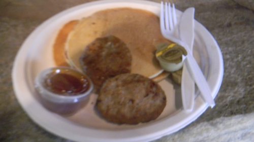 A plate with two sausage patties, a pancake, syrup in a cup, and plastic utensils.