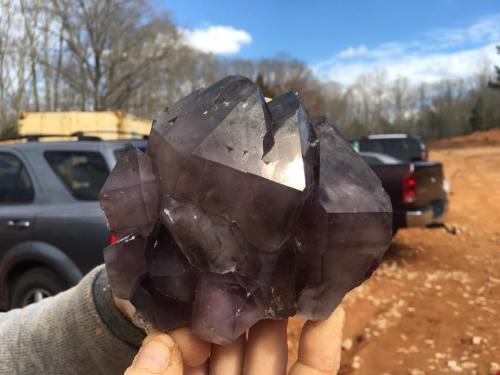 A person holds a large, purple amethyst crystal against a backdrop of trees and blue sky.