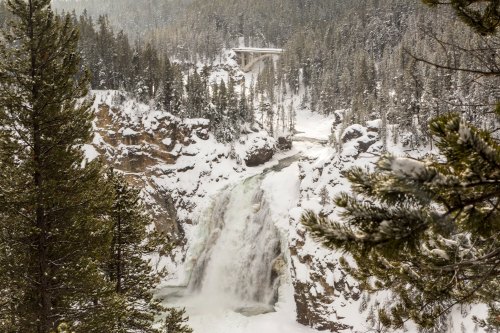 A snowy waterfall cascades down rocky cliffs, surrounded by evergreen trees and a bridge in the background.