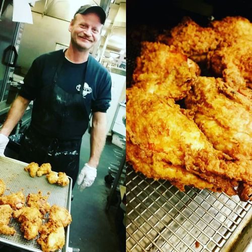 A smiling chef stands next to a tray of freshly fried chicken in a kitchen setting.