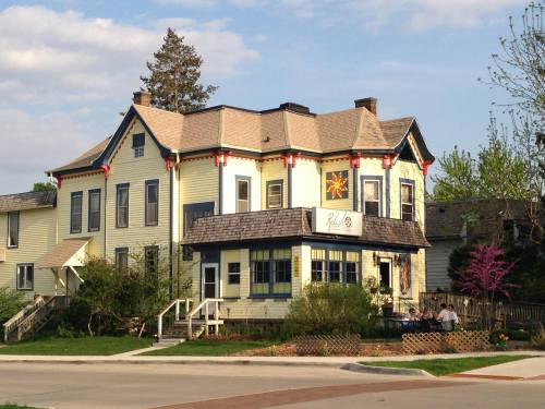 A charming yellow Victorian house with a decorative roof and porch, surrounded by greenery and a clear blue sky.