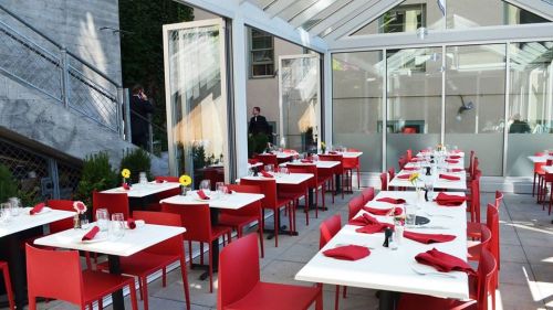 A modern restaurant patio with red chairs and tables set for dining, featuring glass walls and greenery.