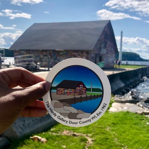 A hand holds a circular sticker of the Hardy Gallery, with a scenic view of the gallery and water in the background.