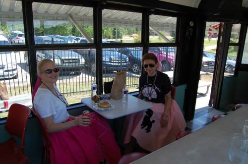 Two women in vintage-style outfits sit at a table inside a diner, enjoying food and drinks, with cars visible outside.