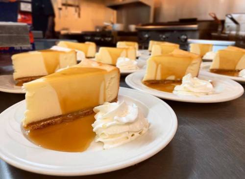 Slices of cheesecake drizzled with caramel sauce, served with whipped cream on white plates in a kitchen setting.