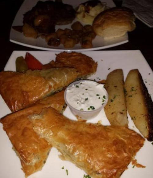A plate featuring flaky pastries, a small cup of dip, and roasted potatoes, with garnishes and a side of pickles.