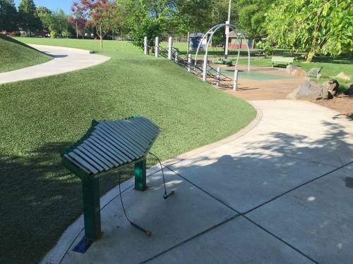 A green xylophone sits on a paved path in a park, surrounded by grass and playground equipment in the background.