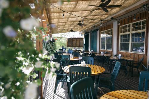 Outdoor dining area with wooden tables and green chairs, shaded by a canopy, surrounded by plants and string lights.