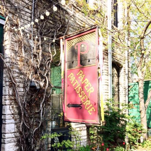 A vintage sign for "Paper Patisserie" hangs outside a charming stone building, surrounded by greenery and string lights.