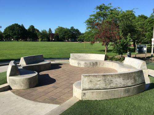 A circular concrete seating area in a park, surrounded by grass and trees under a clear blue sky.