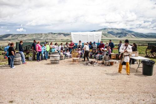 A large group of people gathers outdoors near barrels and wagons, with mountains in the background and a white backdrop.
