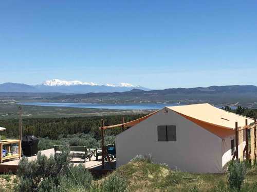 A glamping tent on a hillside with a view of a lake and snow-capped mountains in the background under a clear blue sky.
