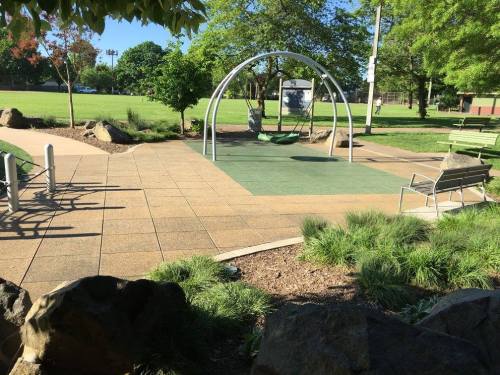 A sunny park scene featuring a swing set, benches, and green grass, surrounded by trees and rocks.