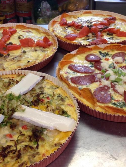 Four savory quiches with various toppings, including vegetables, tomatoes, and slices of sausage, in paper liners.