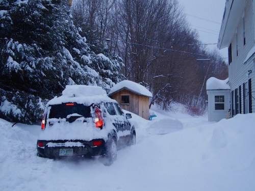 A car covered in snow navigates a snowy driveway beside a house and a small shed in a winter landscape.
