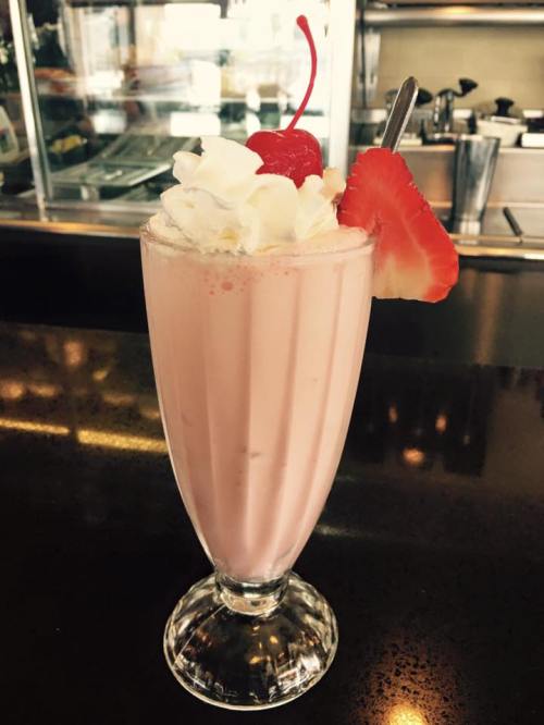 A tall glass of pink milkshake topped with whipped cream, a cherry, and a slice of strawberry.