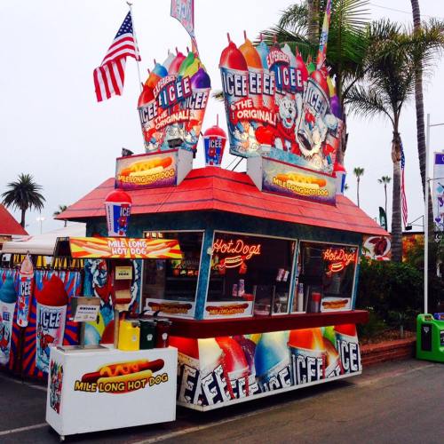 Colorful food stand featuring icee drinks and hot dogs, with flags and vibrant signage in a festive outdoor setting.