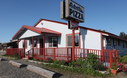 Exterior of Roscoe's Pizza, a white building with red trim and a sign, surrounded by greenery and gravel.