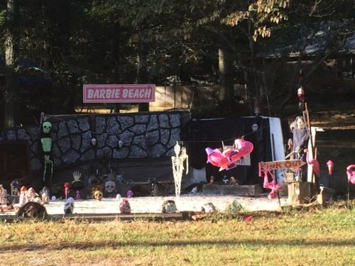 A whimsical Halloween display featuring a "Barbie Beach" sign, skeletons, skulls, and pink flamingos in a sandy area.