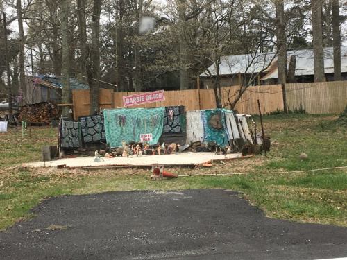 A whimsical outdoor display featuring a "Barbie Beach" sign, decorated with dolls and colorful fabrics in a wooded area.