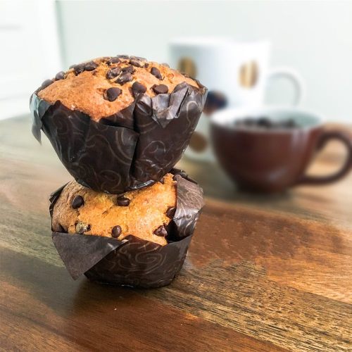 Two chocolate chip muffins stacked on a wooden table, with a coffee cup in the background.