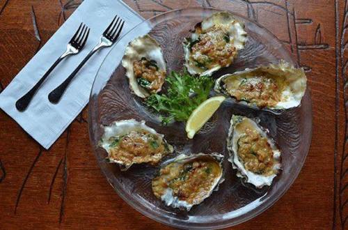 A plate of baked oysters topped with herbs, served with a lemon wedge and garnished with lettuce, alongside two forks.