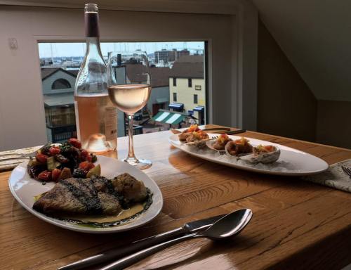 A table set with two plates of seafood dishes, a glass of rosé wine, and a view of a marina through a window.
