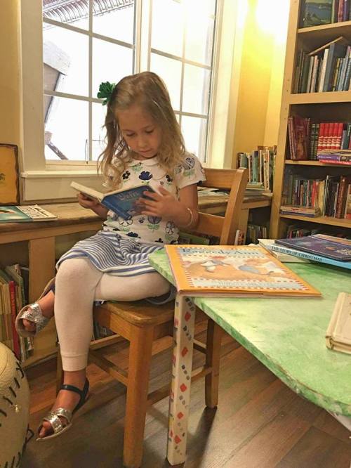 A young girl sits on a chair, reading a book in a cozy library filled with books and natural light.